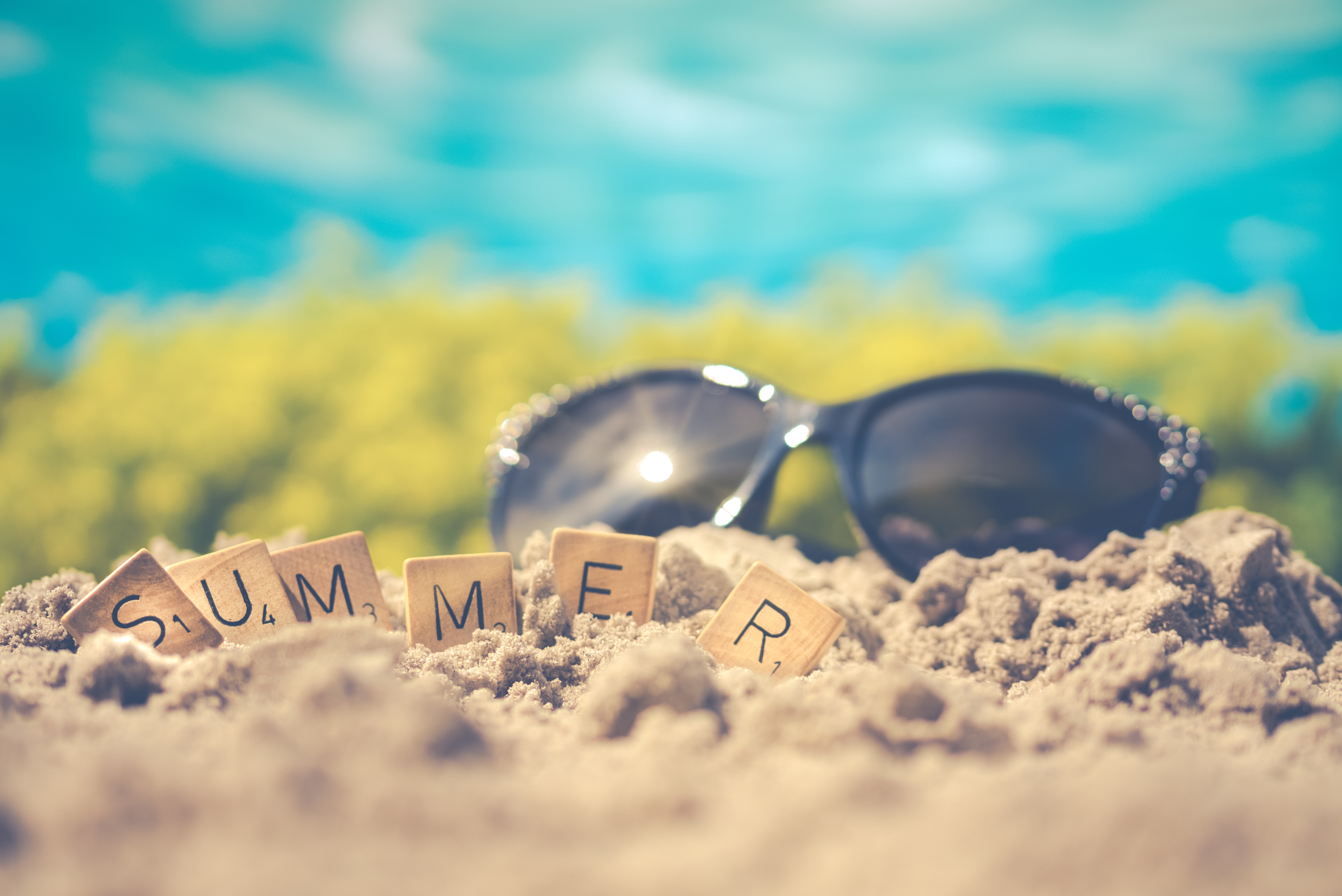 The word summer spelled out with Scrabble letters, sitting in pile of sand with sunglasses in the background