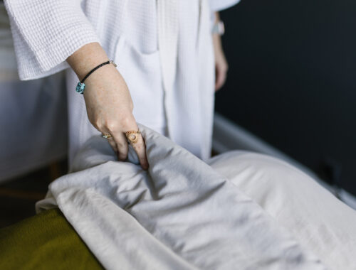 Woman in a spa robe gently pulls back soft white sheets, preparing to receive a peaceful spa treatment.